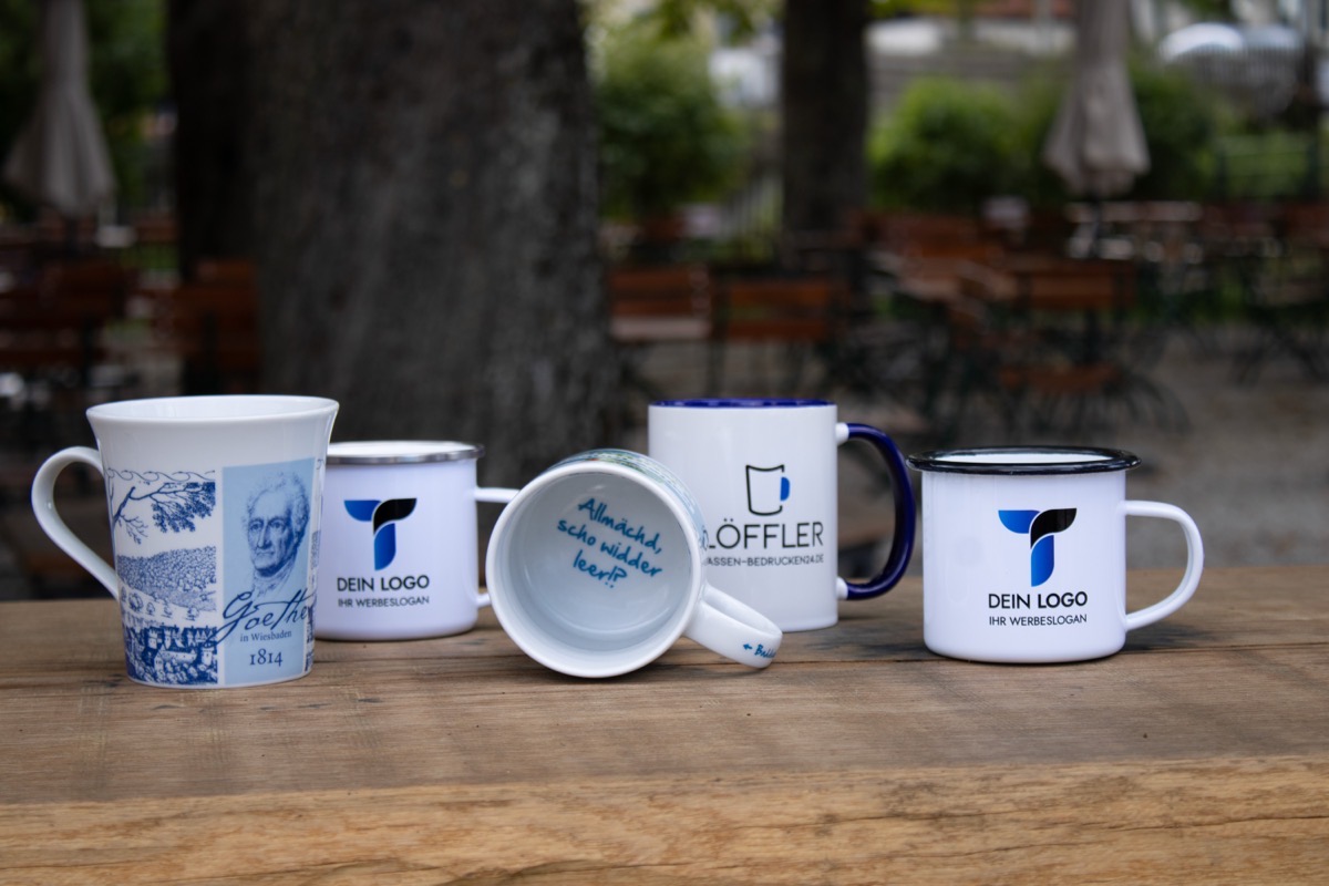 Auswahl bedruckter Tassen mit Logodruck: Emailletassen, Porzellantasse und Keramiktasse auf einem Holztisch